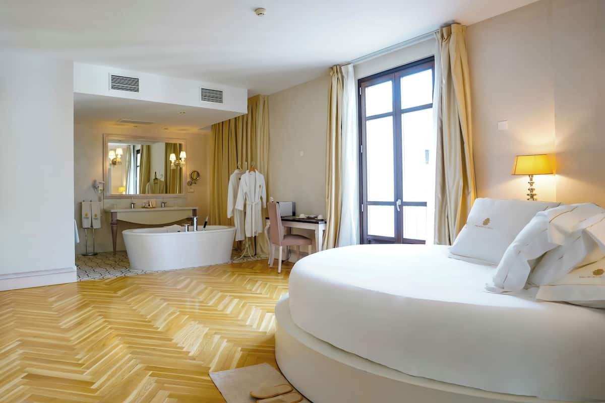 Habitación de hotel con una cama grande, zona de estar con una silla, escritorio, bañera y un ventanal con cortinas.