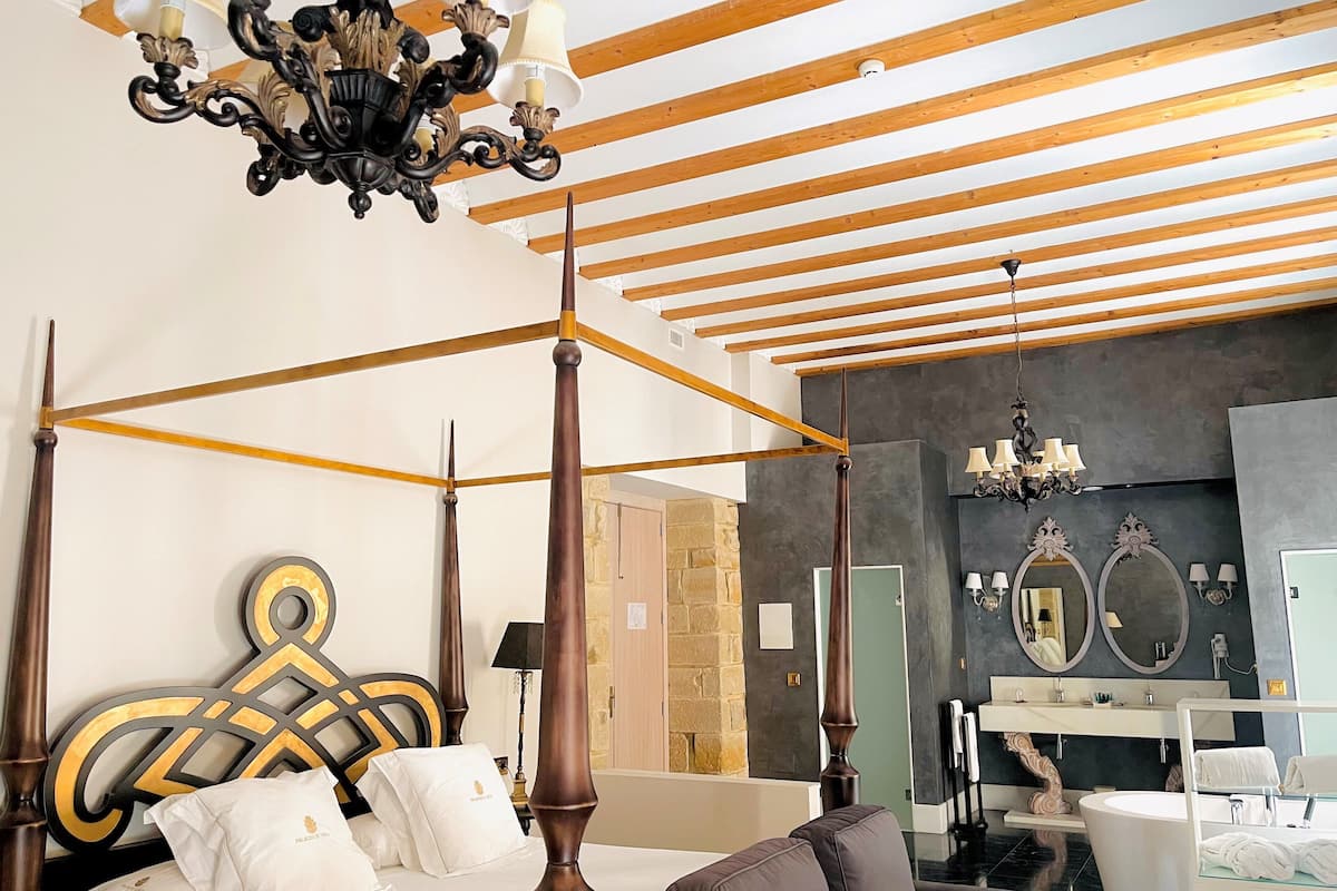 Una cama con dosel, cabecera de madera, candelabro y un baño moderno con un espejo grande y bañera.
