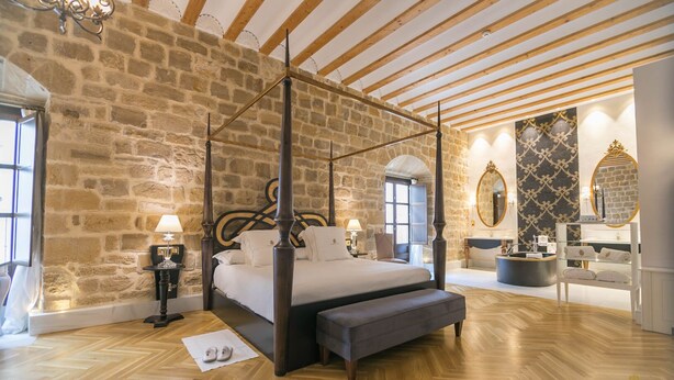 A bedroom with a four-poster bed, stone walls, wooden beams, a vanity with a mirror, and a reception desk.