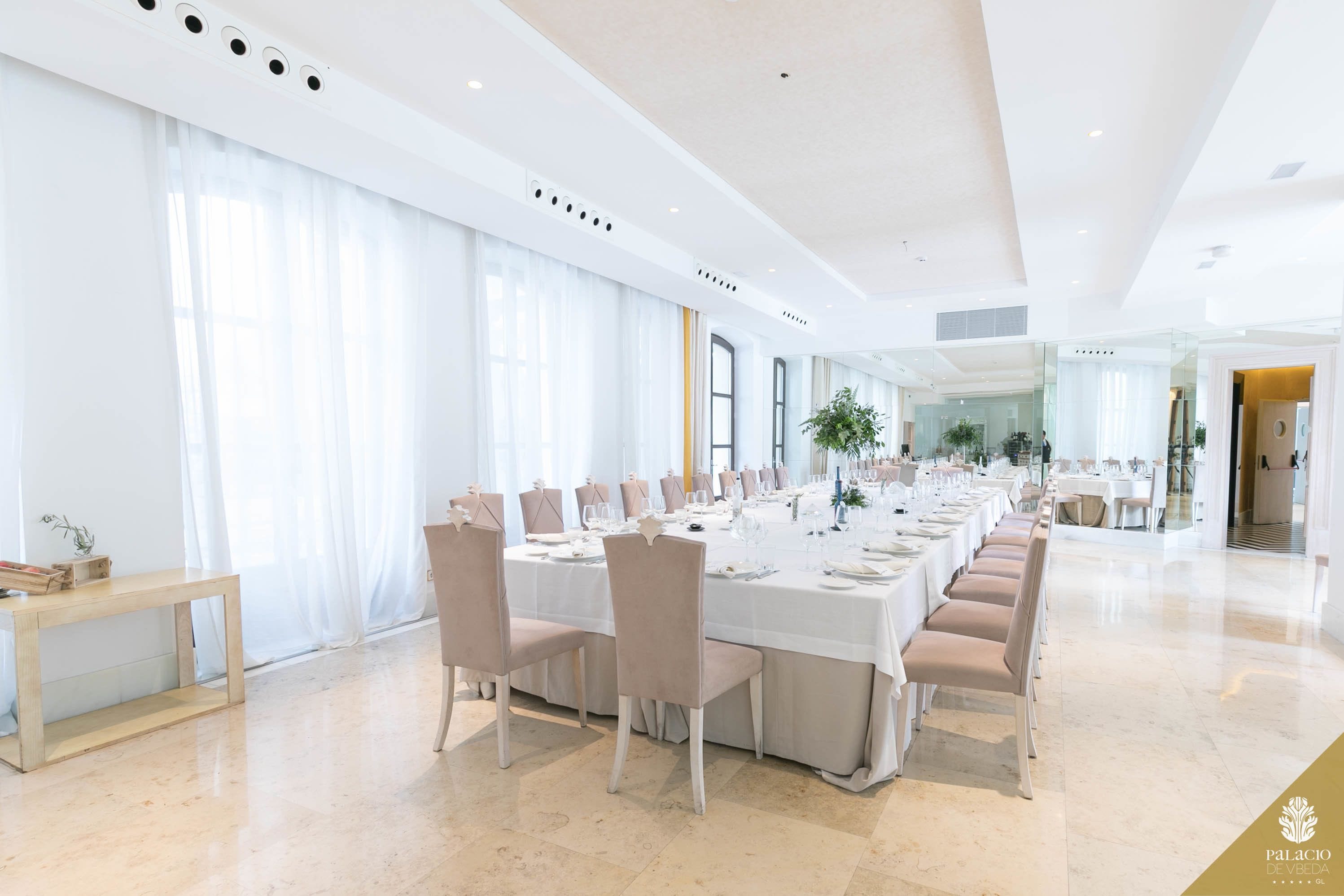 Un vaste espace dédié à la salle à manger, avec de longues tables dressées pour le repas, de grandes fenêtres ornées de rideaux légers et un aménagement intérieur épuré et moderne.