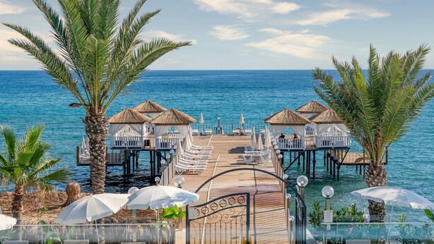 Un muelle de madera que lleva a bungalows sobre el agua con techo de paja, palmeras y vistas al mar.