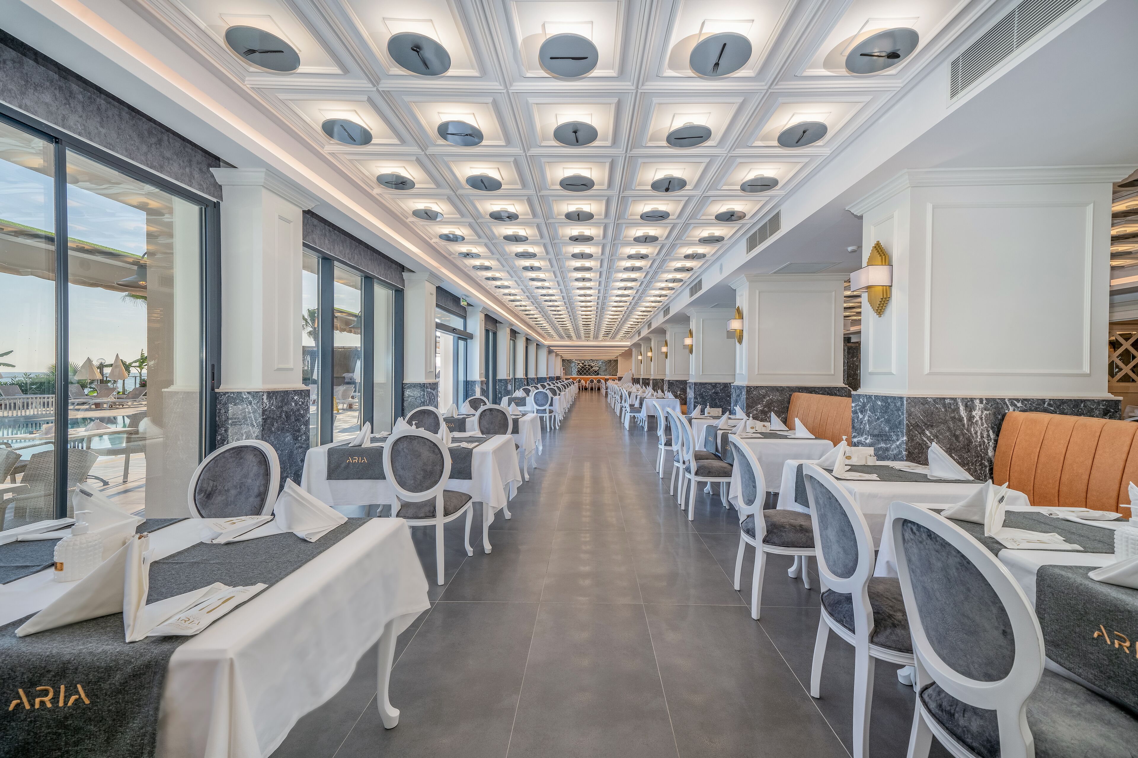 A restaurant with a long, central aisle, white tables and chairs, and a ceiling with circular lights.