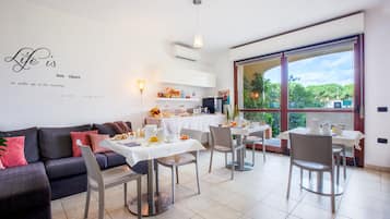 Ein stilvolles Wohnzimmer mit Sofa, Esstisch und Stühlen. Eine Küchenzeile mit Kühlschrank, Mikrowelle und verschiedenen Küchenutensilien ist ebenfalls vorhanden. Ein großes Fenster bietet Blick ins Grüne.