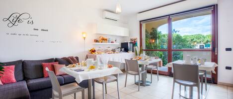 Una sala de estar moderna con sofá, mesa de comedor y sillas. Hay una zona de cocina con refrigerador, microondas y diversos utensilios. Un ventanal ofrece vistas al exterior con vegetación.
