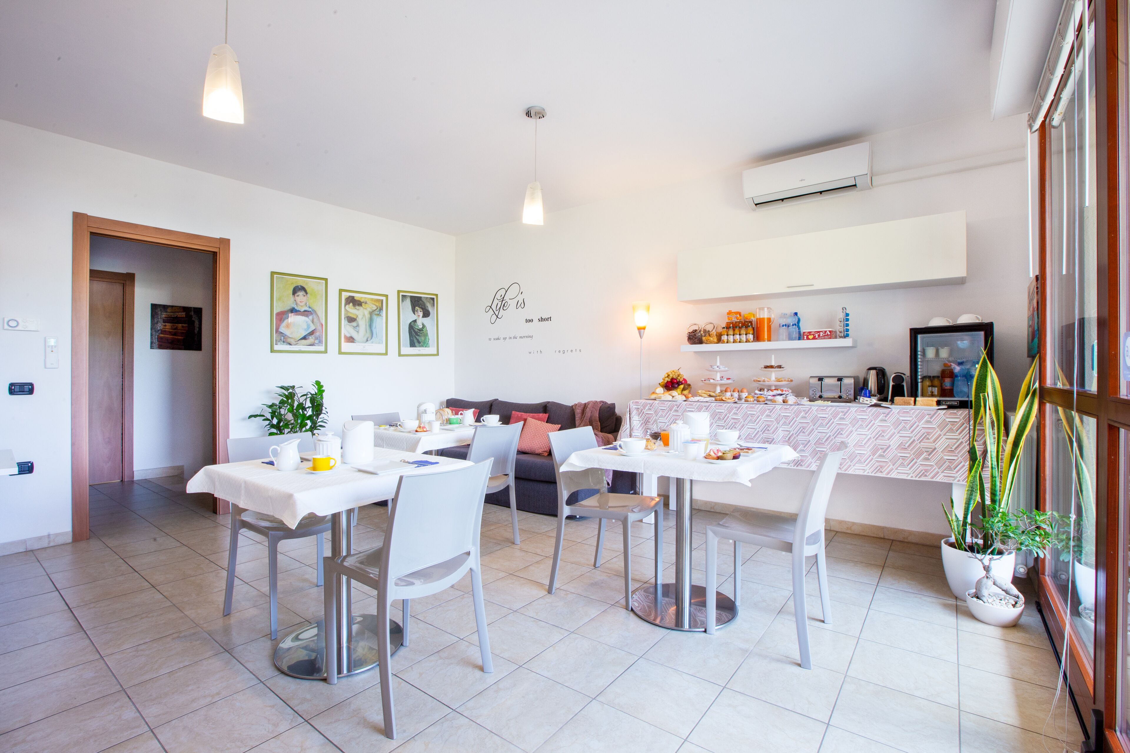 Un comedor moderno con mesas y sillas blancas, una cocina al fondo y una pared con cuadros y una cita.