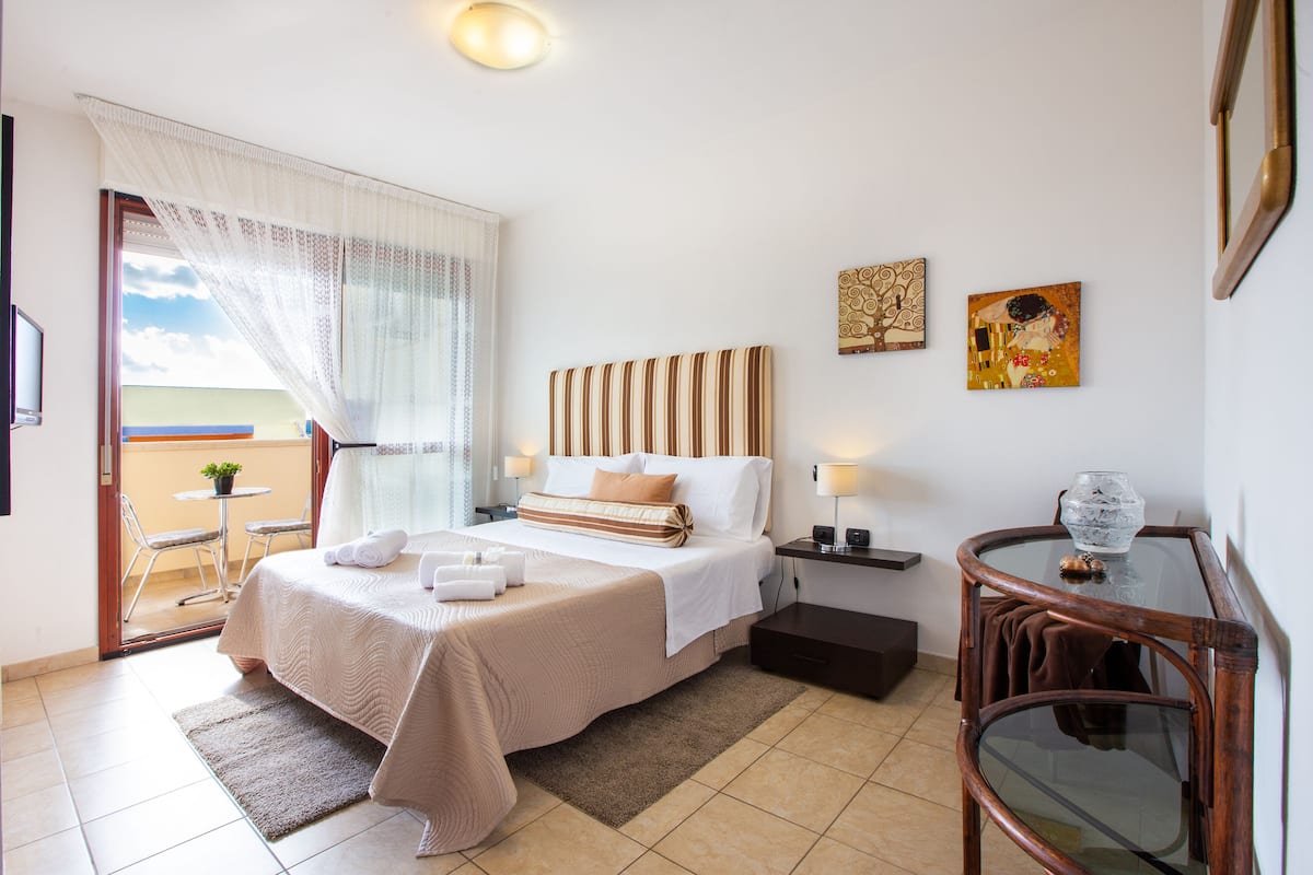 A hotel room with a bed, a nightstand, a glass table, and a view of the outdoors.