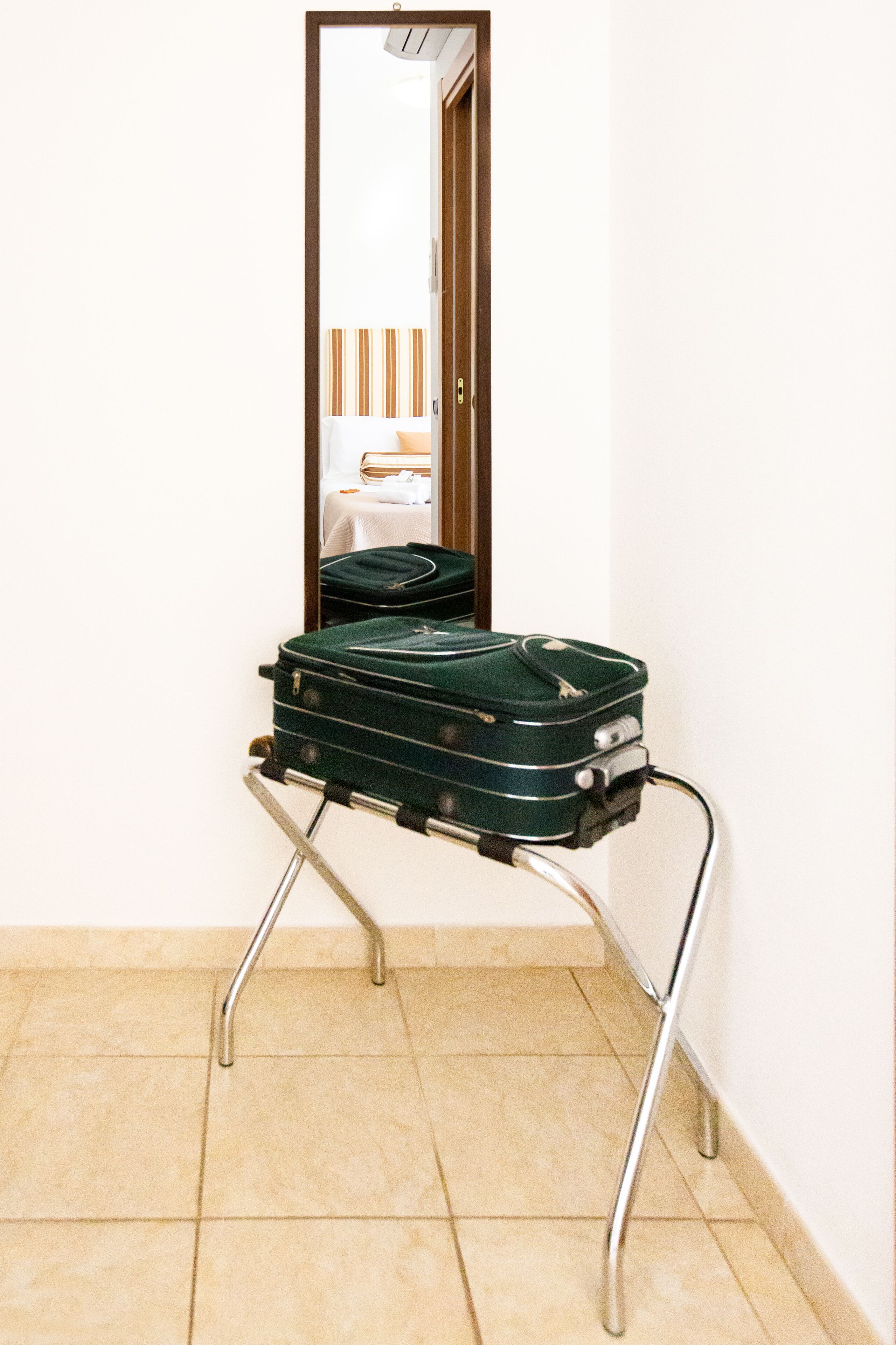 A green suitcase on a wheeled stand in a room with a tiled floor and a mirror reflecting a bed and a window.