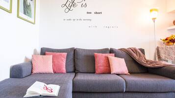 En grå sofa med pink og brune puder, et indramet citat på væggen og et natbord med lampe og skål med frugt.