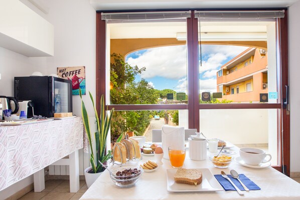 Una mesa de desayuno con diferentes alimentos, una cafetera y vista a un edificio a través de una ventana.