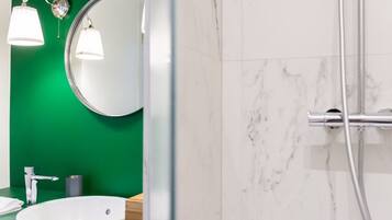 Un baño con pared verde, un espejo redondo, un lavamanos blanco y una zona de ducha con pared de mármol.