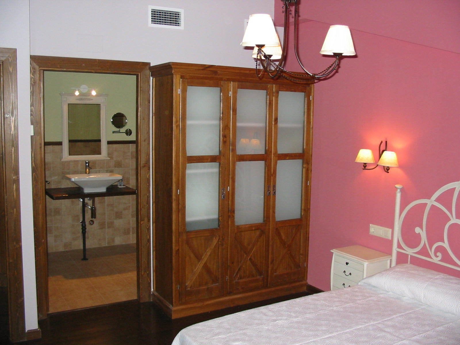 Quarto com guarda-roupa de madeira, mesa de cabeceira, cama, espelho e banheiro com pia e espelho.
