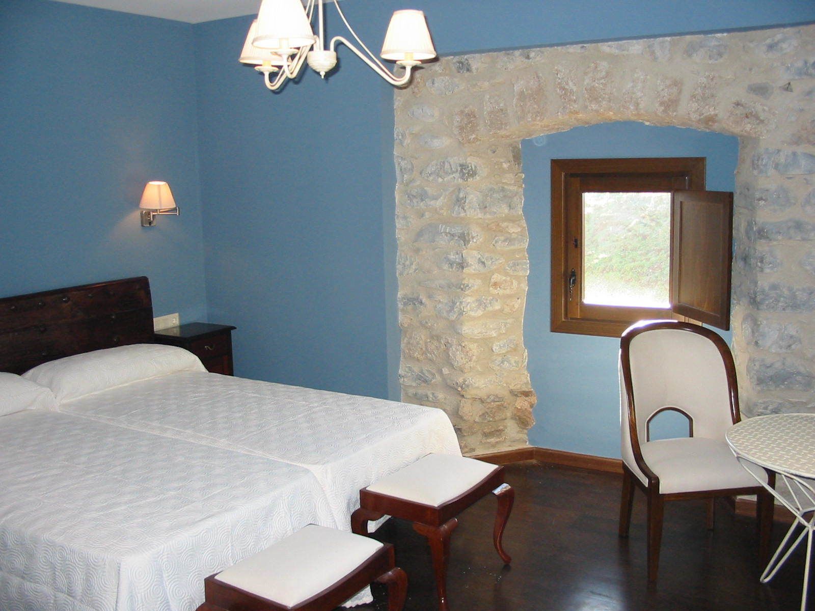A bedroom with a large bed, a stone wall, a window, a chair, and a table.