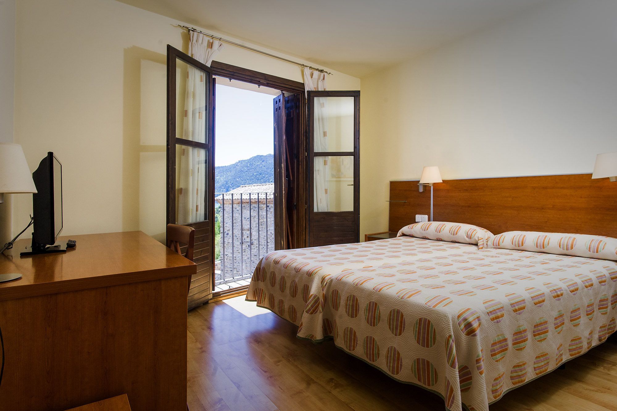 Hotelkamer met een houten bureau, een bed met gestreepte beddengoed, een nachtkastje met een lamp en een balkon met uitzicht op de bergen.