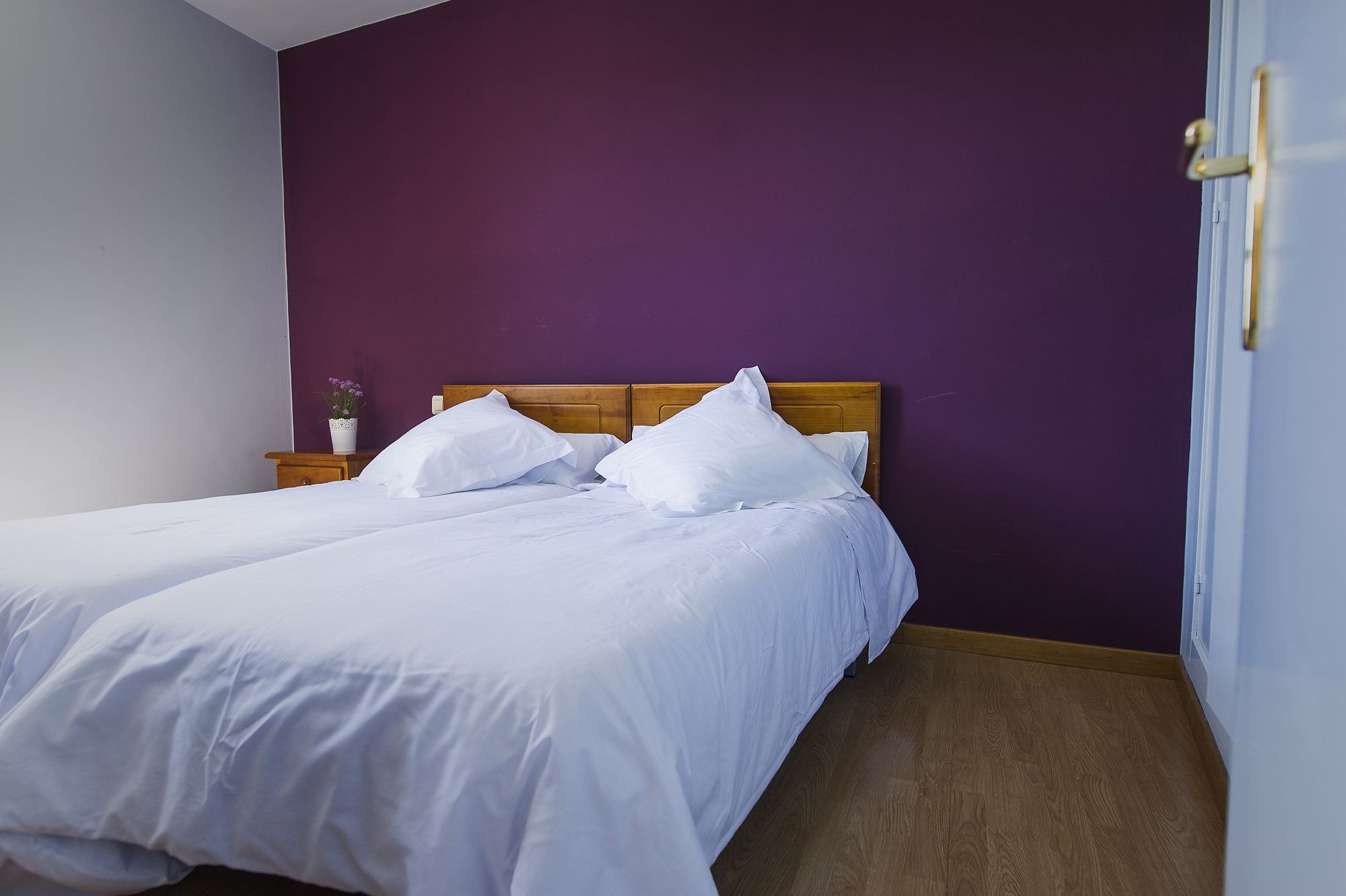 Un lit bien fait, avec du linge de lit blanc, dans une pièce aux murs violets et au plancher en bois.