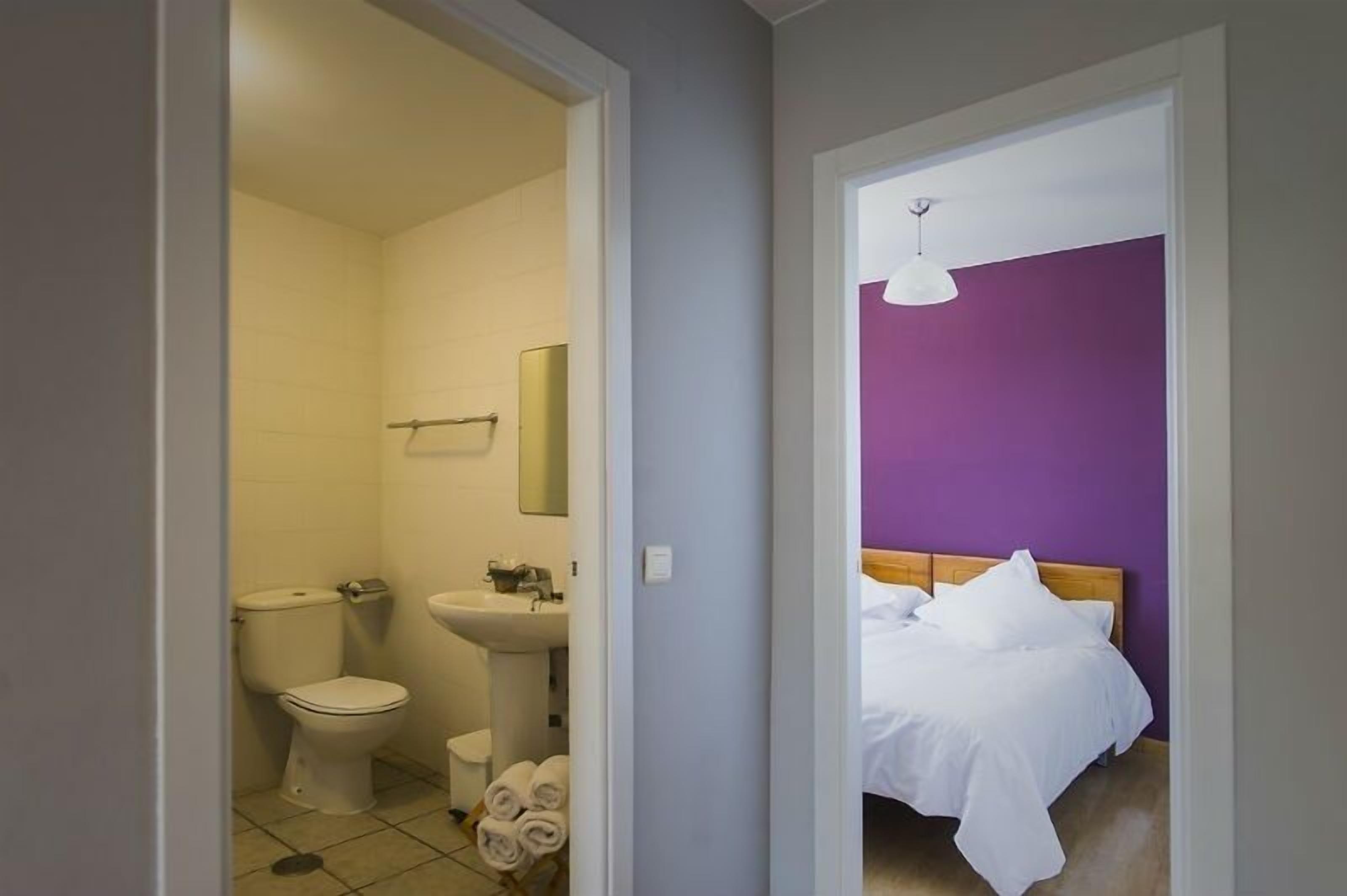 Ett hotellrum med ett badrum som innehåller en toalett, ett handfat och en spegel. Rummet har en säng med vita sängkläder och en lila vägg.