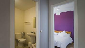 Ett hotellrum med ett badrum som innehåller en toalett, ett handfat och en spegel. Rummet har en säng med vita sängkläder och en lila vägg.