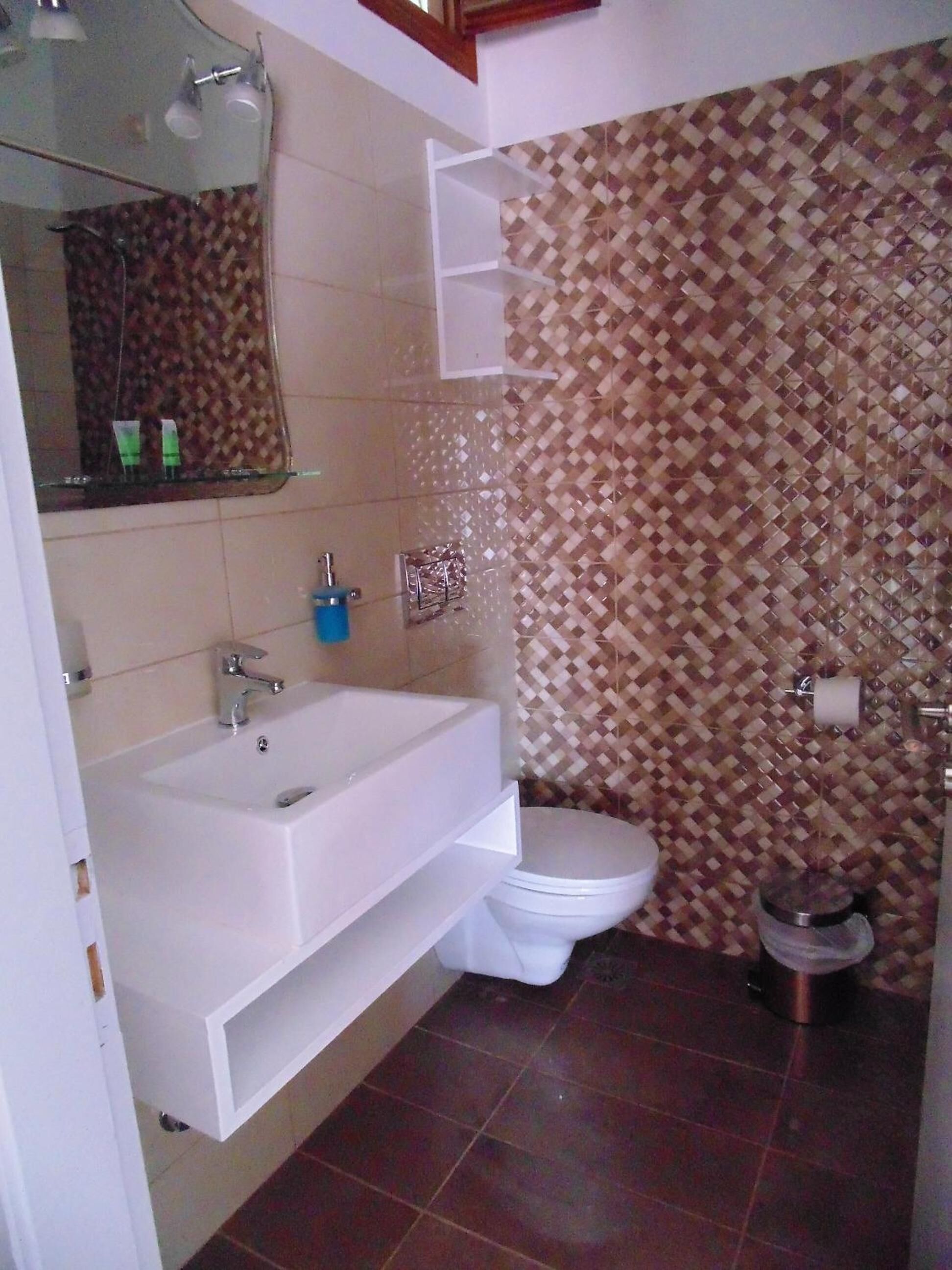 A bathroom with a white sink, mirror, and toilet. There is a patterned tile wall and a small shelf above the sink.