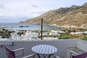 A balcony with a table and chairs overlooking a coastal town with white buildings and a ship in the distance.