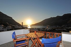 A wooden table and chairs on a balcony overlooking a coastal town at sunset.