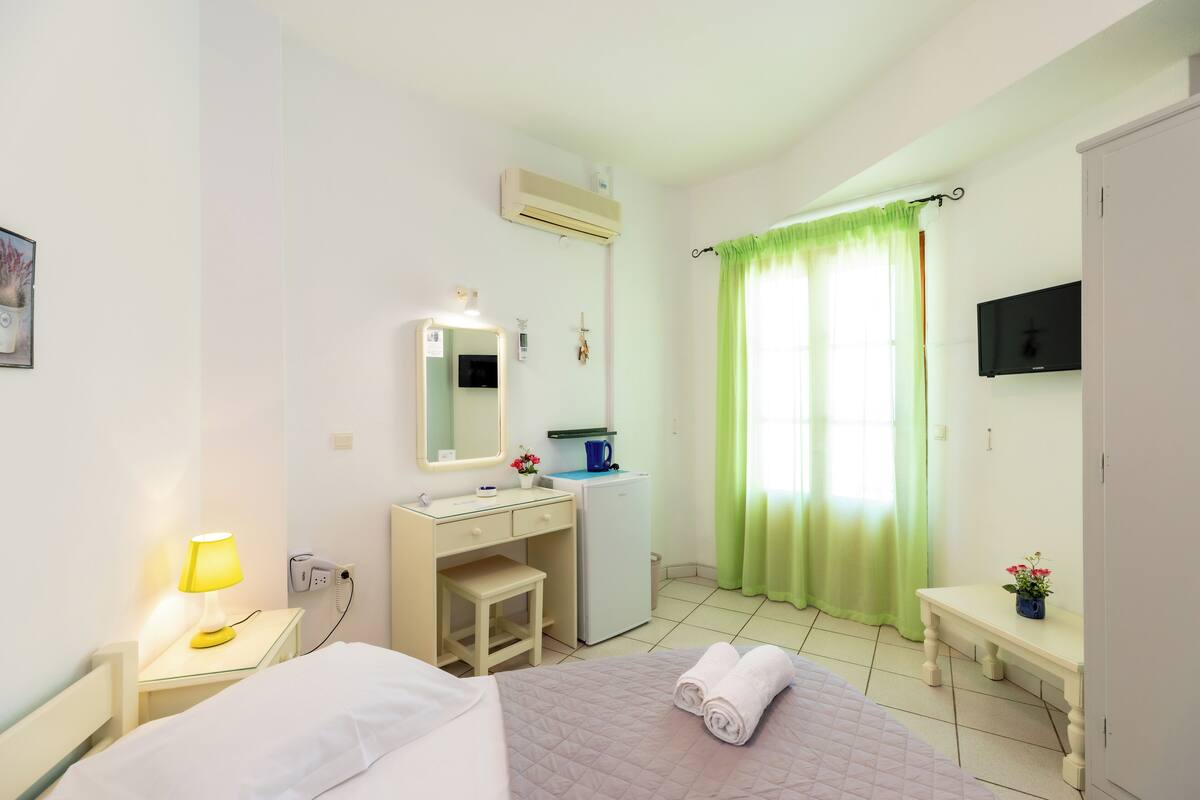 A hotel room with a bed, bedside table with a lamp, a desk with a mirror, a small stool, a refrigerator, a TV mounted on the wall, and a window with green curtains.