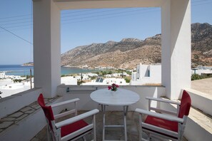 A balcony with a white table and two red chairs, overlooking a coastal town and mountains.