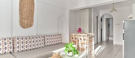 A modern living room with a sofa, a dining table with chairs, a kitchen area, and a hallway.