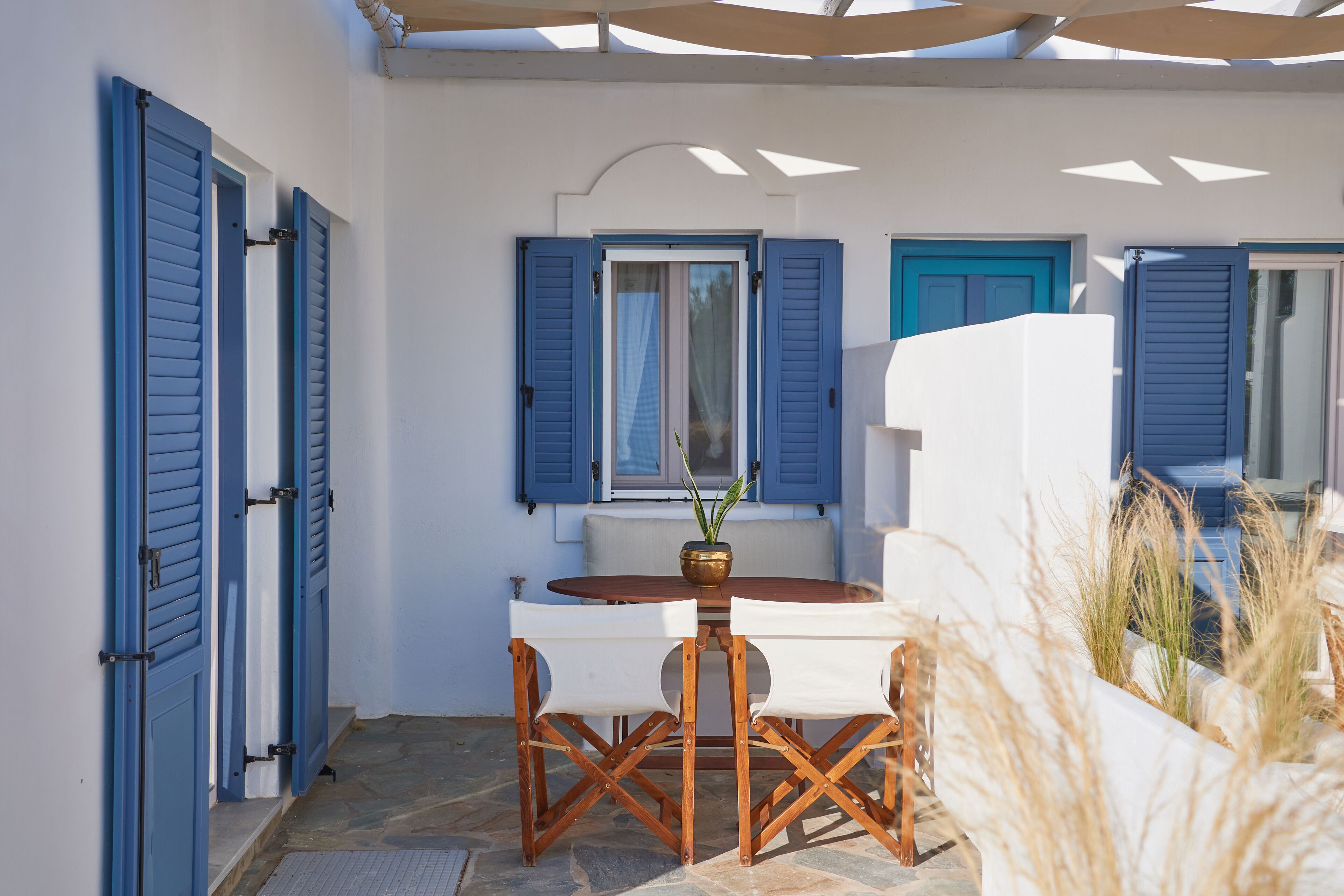 A covered outdoor seating area with a table and chairs, a potted plant, and a view of a building with blue shutters.
