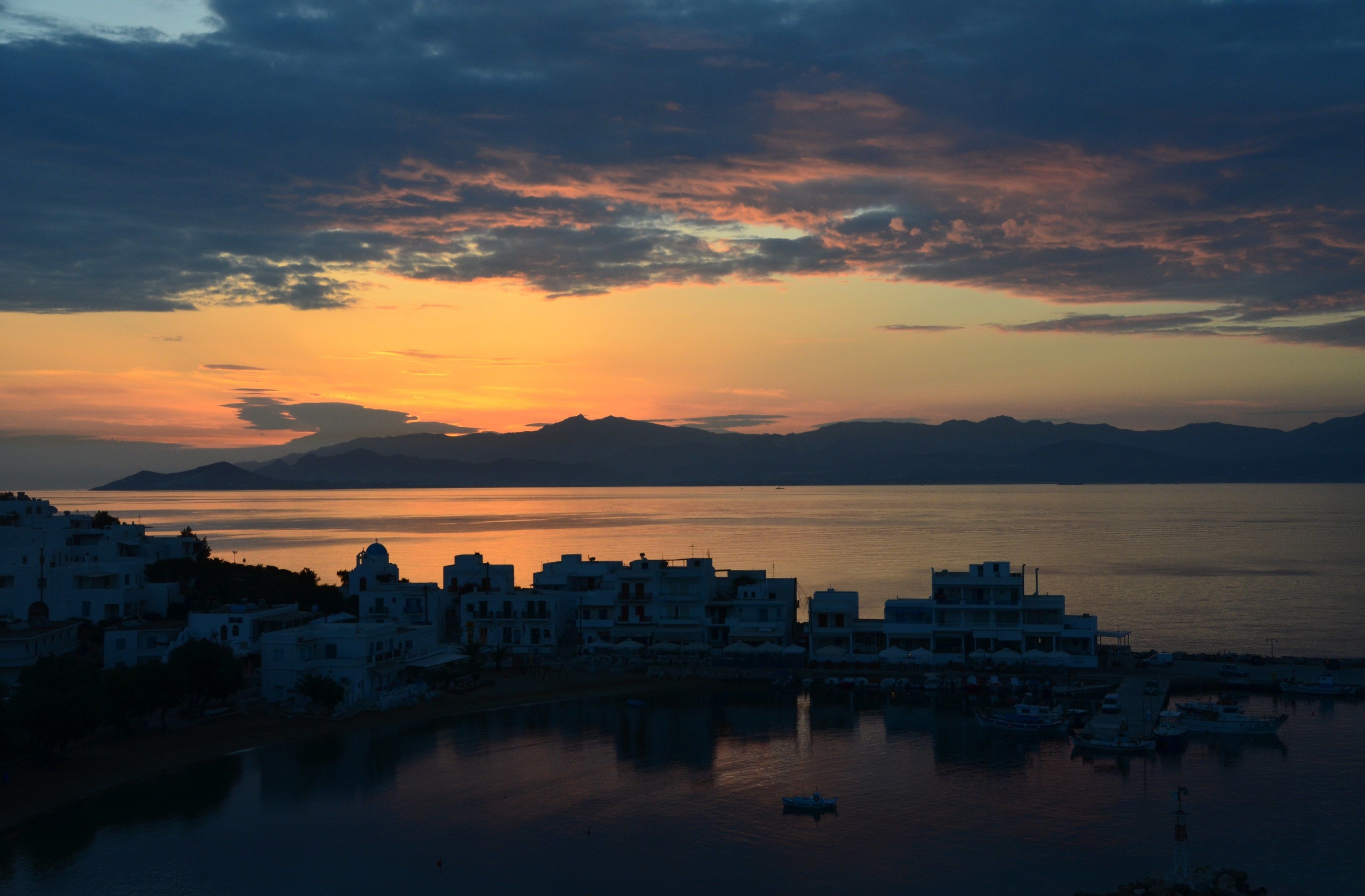 A coastal town with buildings along the waterfront, a marina with boats, and a sunset over the sea.