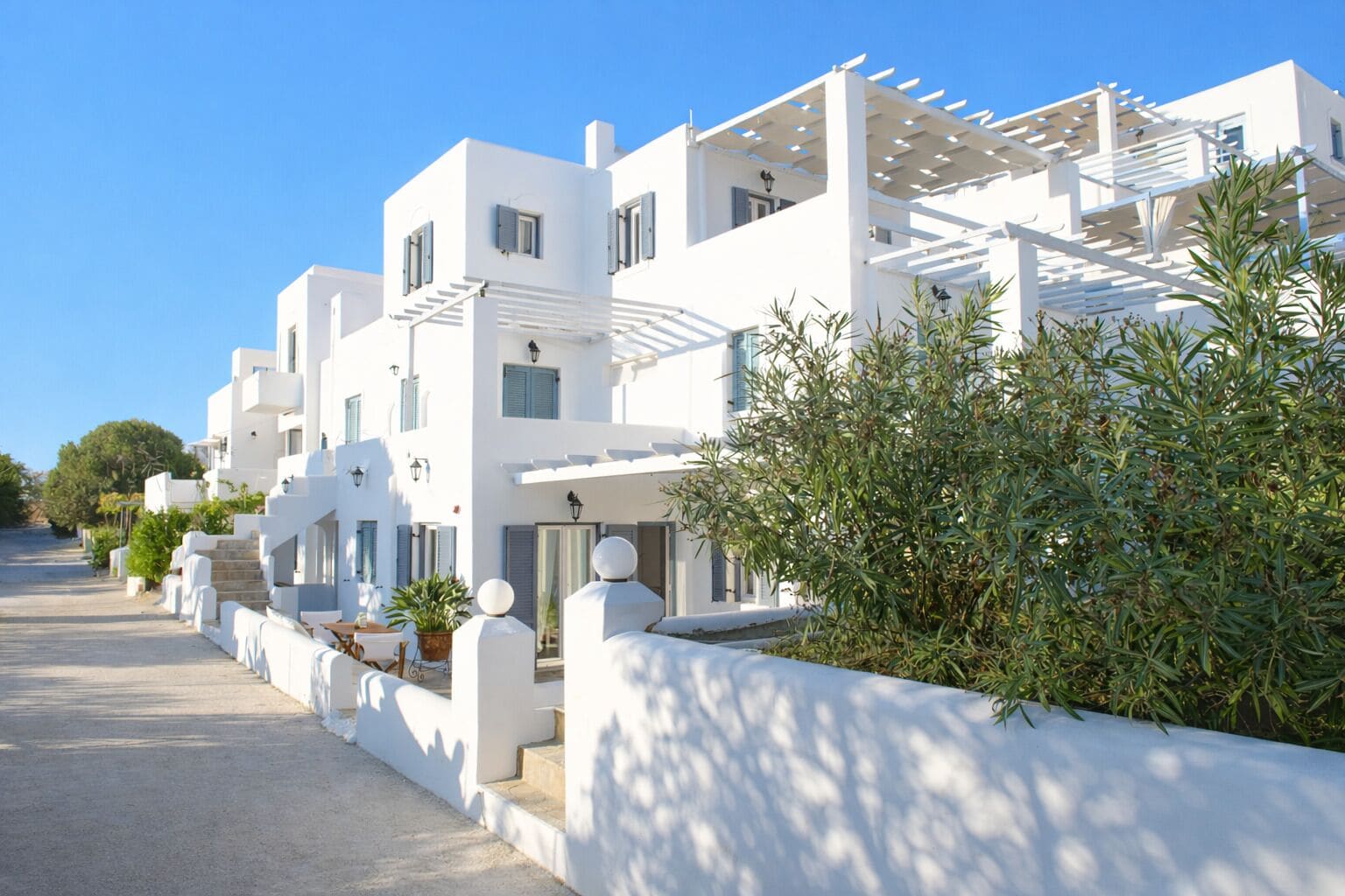 Una hilera de edificios blancos con balcones, un paseo empedrado y algo de vegetación.