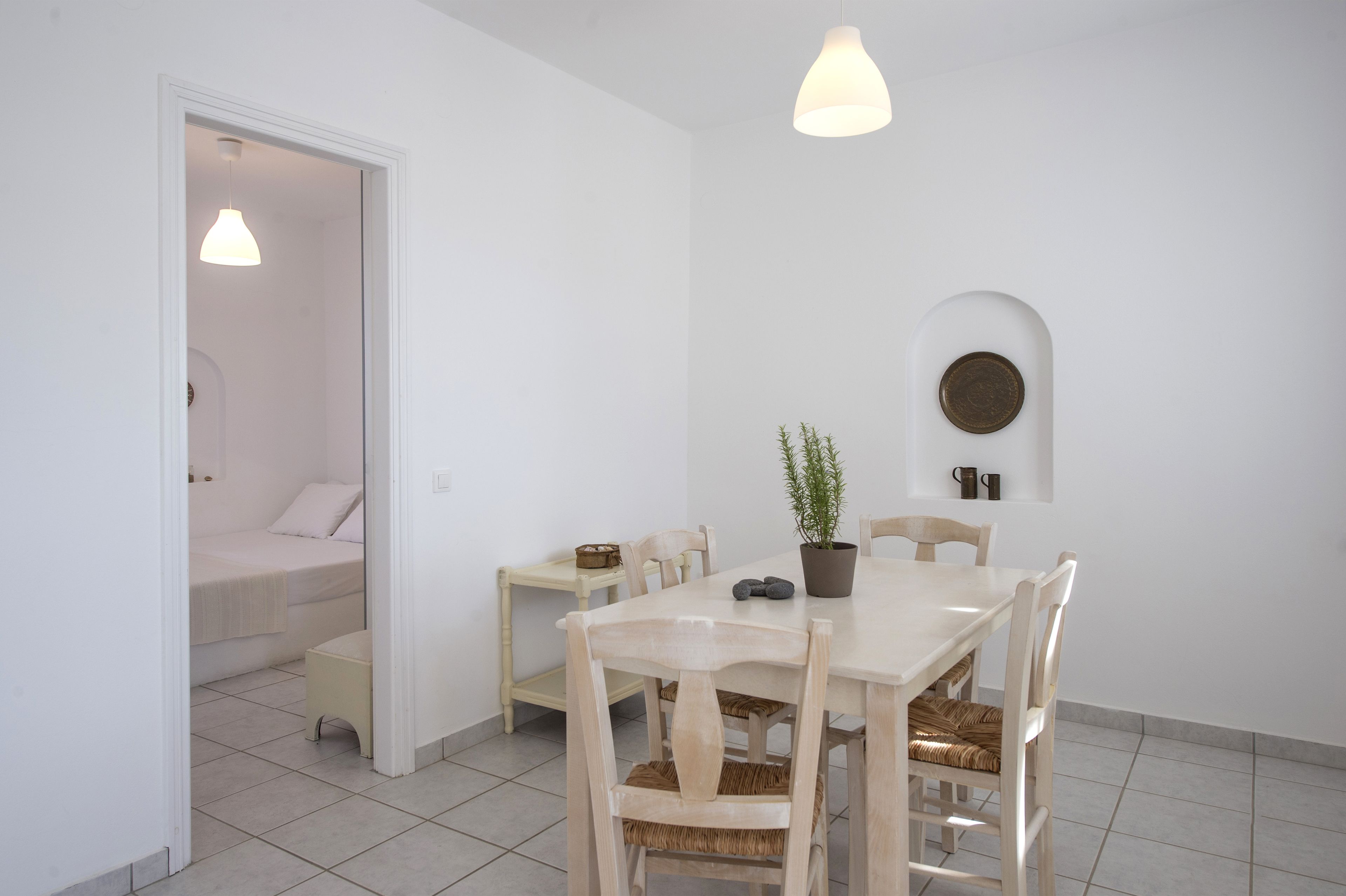 A minimalist dining area with a white table, wooden chairs, a potted plant, and a wall-mounted light fixture.