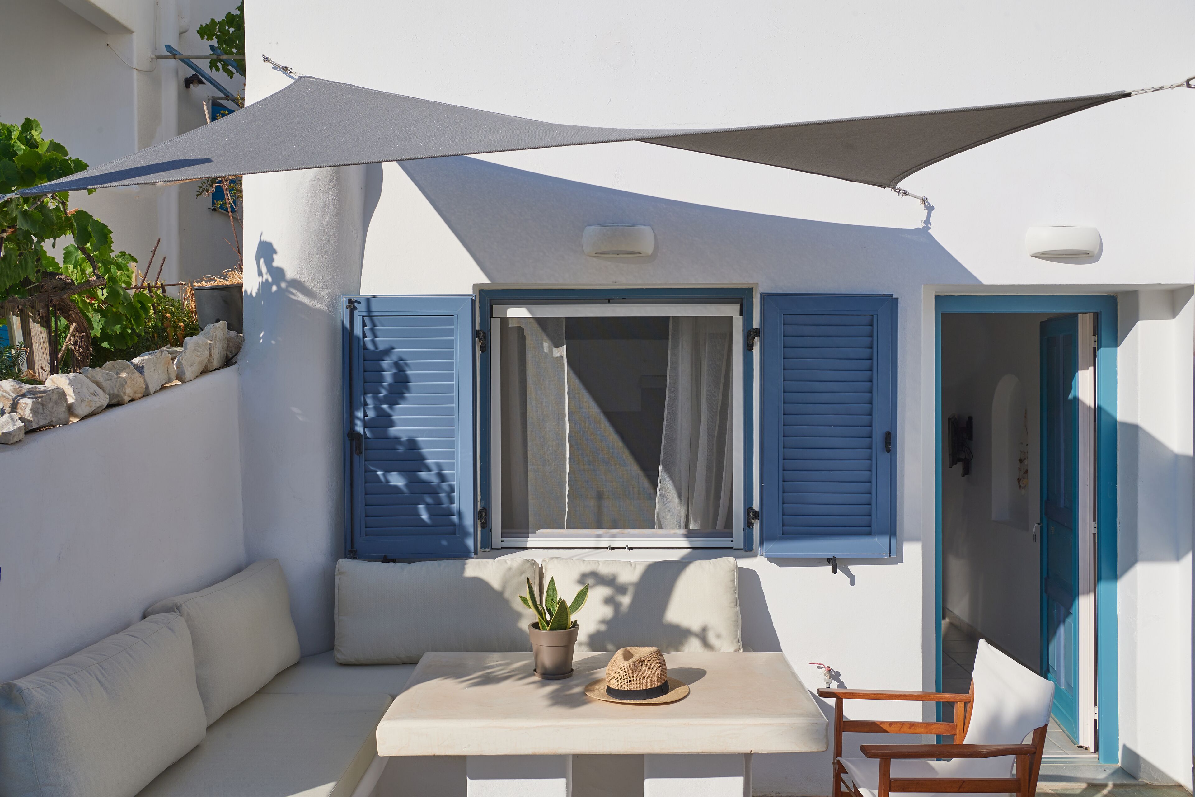 A white building with blue shutters, a table with a potted plant and a hat, and a wooden chair.