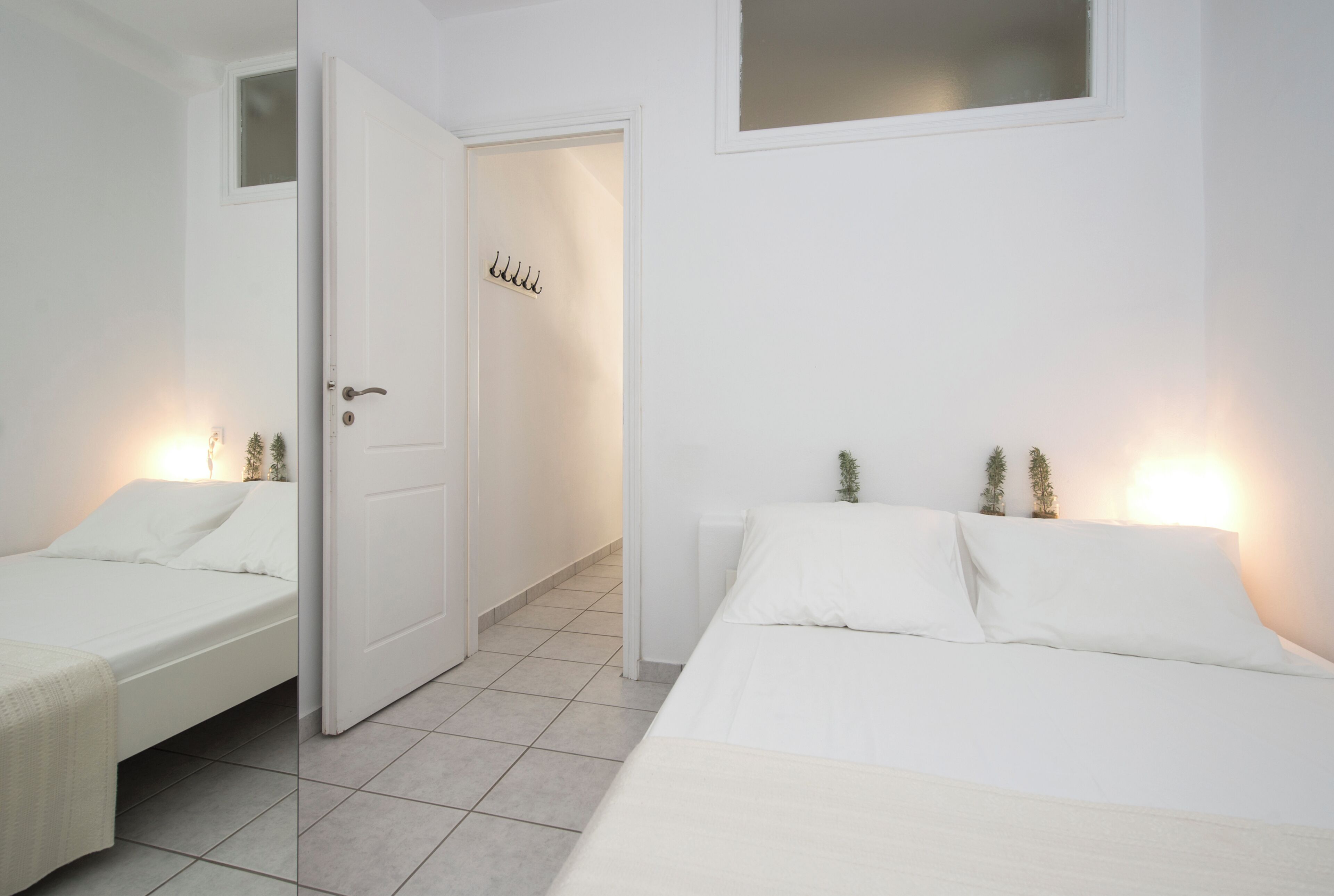 Quarto minimalista com duas camas, uma porta branca e uma janela pequena.