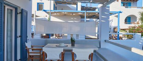 A covered outdoor seating area with a table and chairs, a potted plant, and a view of a white building with blue accents.
