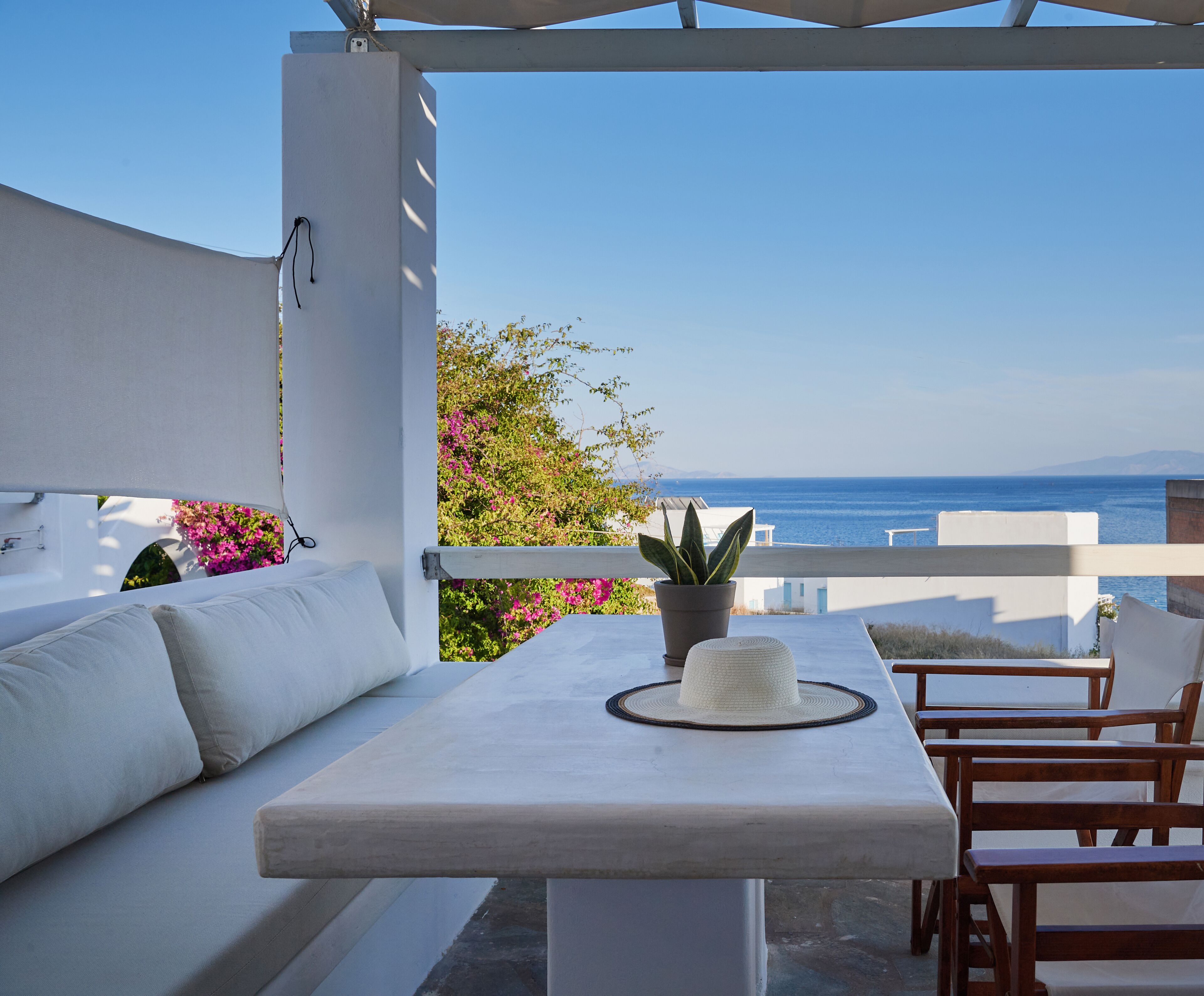 A terrace with a white sofa, a table, and wooden chairs. There is a potted plant on the table. The terrace overlooks a body of water and a distant landmass.