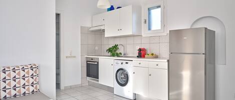 A modern kitchen with white cabinetry, a stainless steel refrigerator, a washing machine, and a dining area with a patterned cushion.