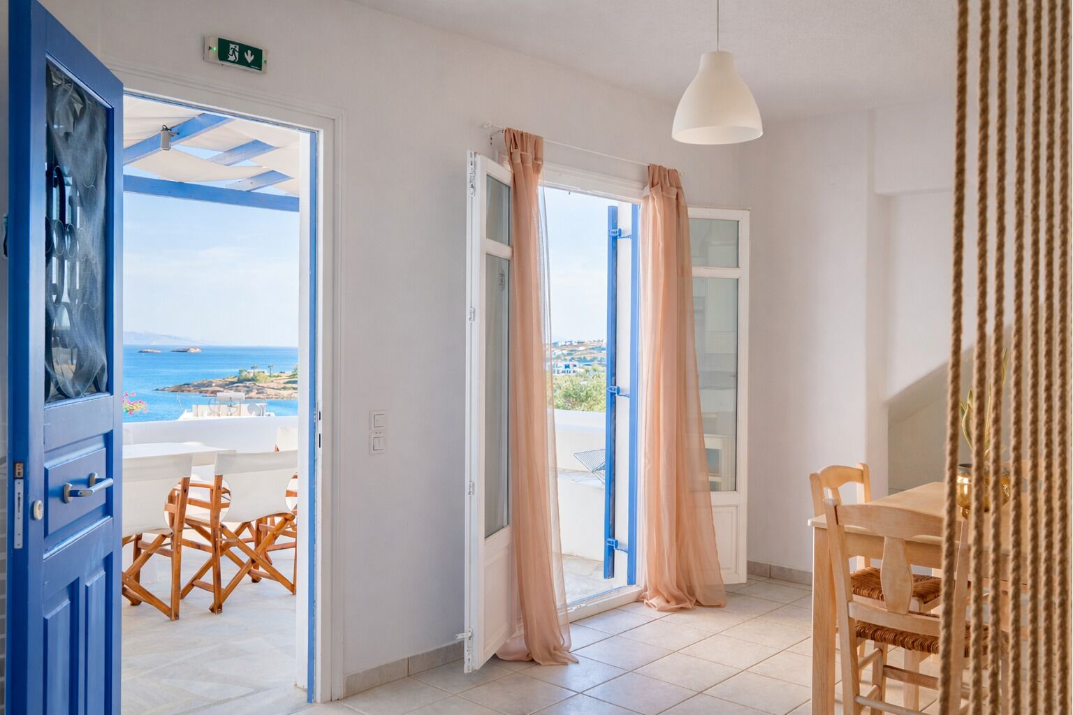 A room with a view of the sea, a table, chairs, and a hanging light fixture.