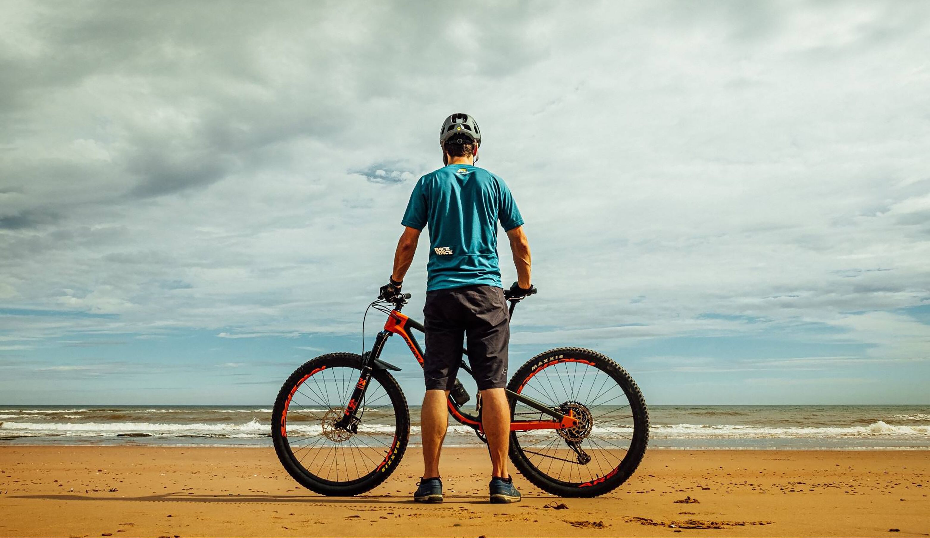 En person står på en sandstrand og holder en mountainbike, med oceanen og en overskyet himmel i baggrunden.