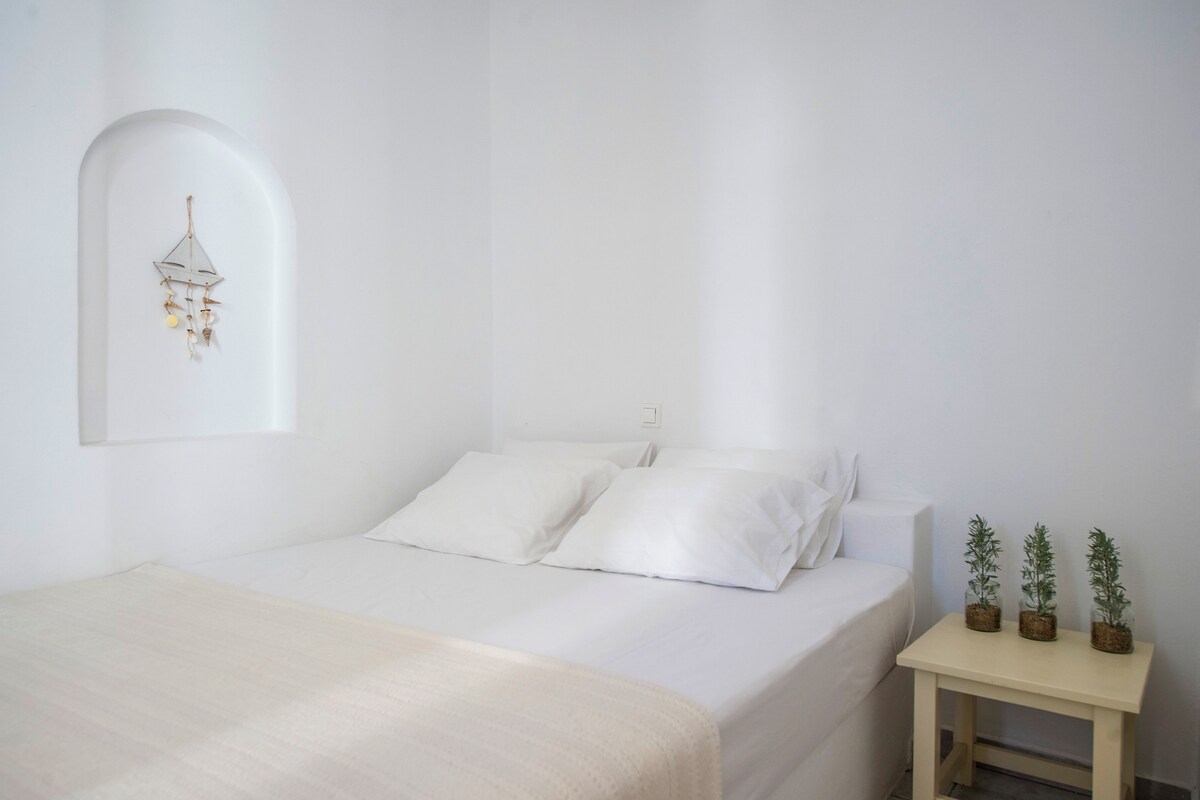 A minimalist bedroom with a bed, white walls, a small bedside table with potted plants, and a wall-mounted light fixture.