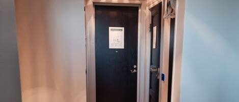 A hotel hallway with a wooden floor, a trash bin, a bed with a blanket, and a door with a sign.