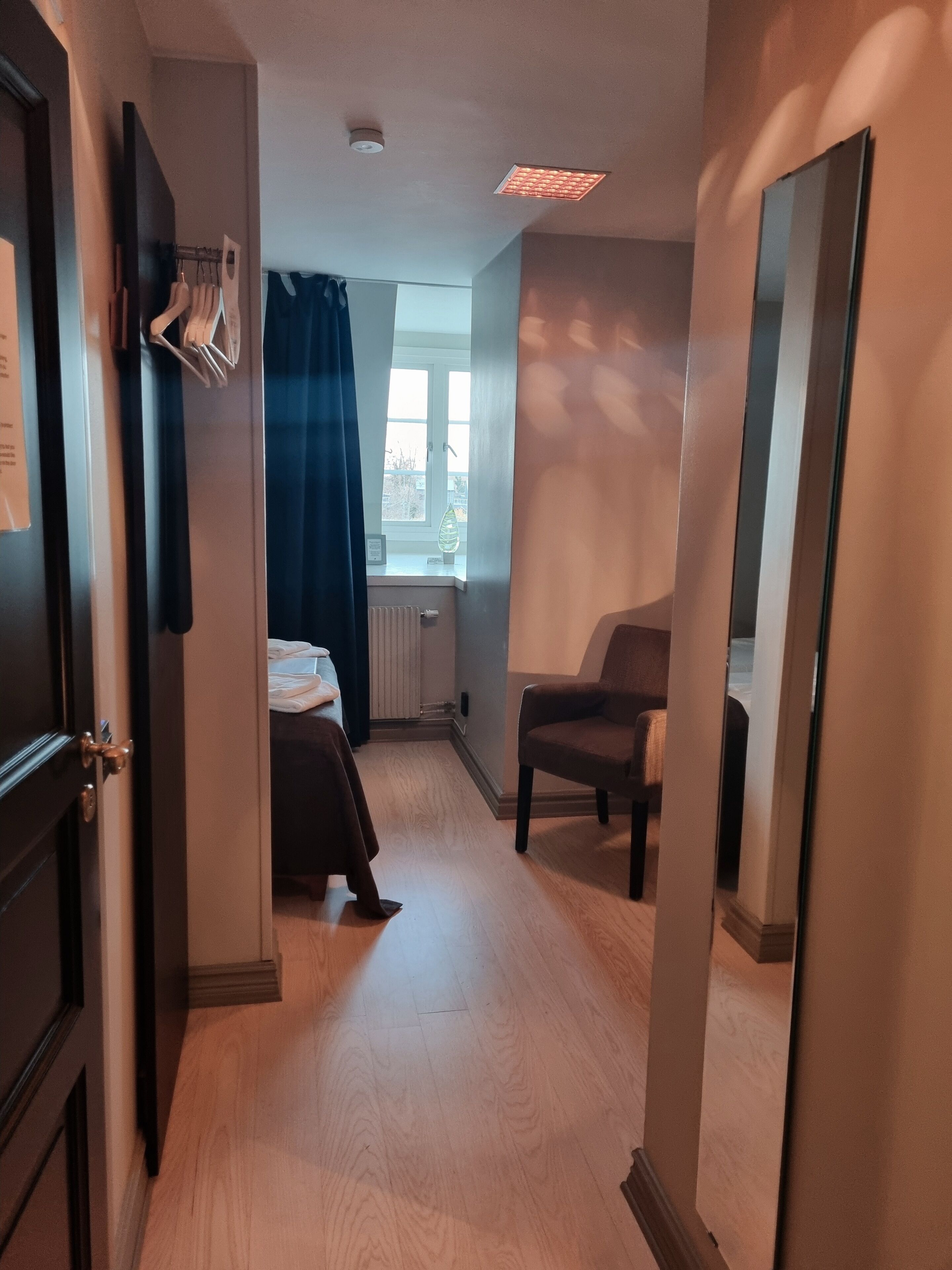 A hotel room with a wooden floor, a bed, a chair, a desk, and a window with curtains.