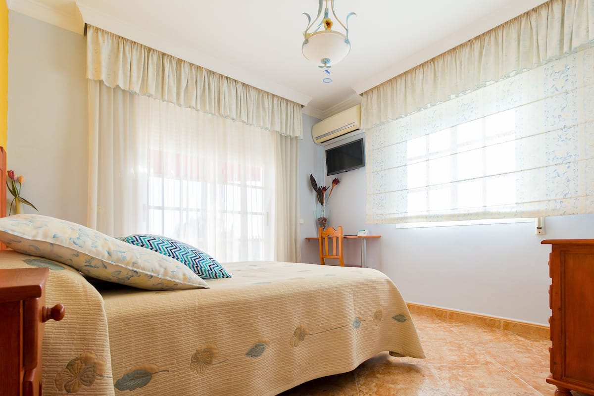 A bedroom with a bed, a nightstand, a wooden dresser, a TV mounted on the wall, and a window with curtains.