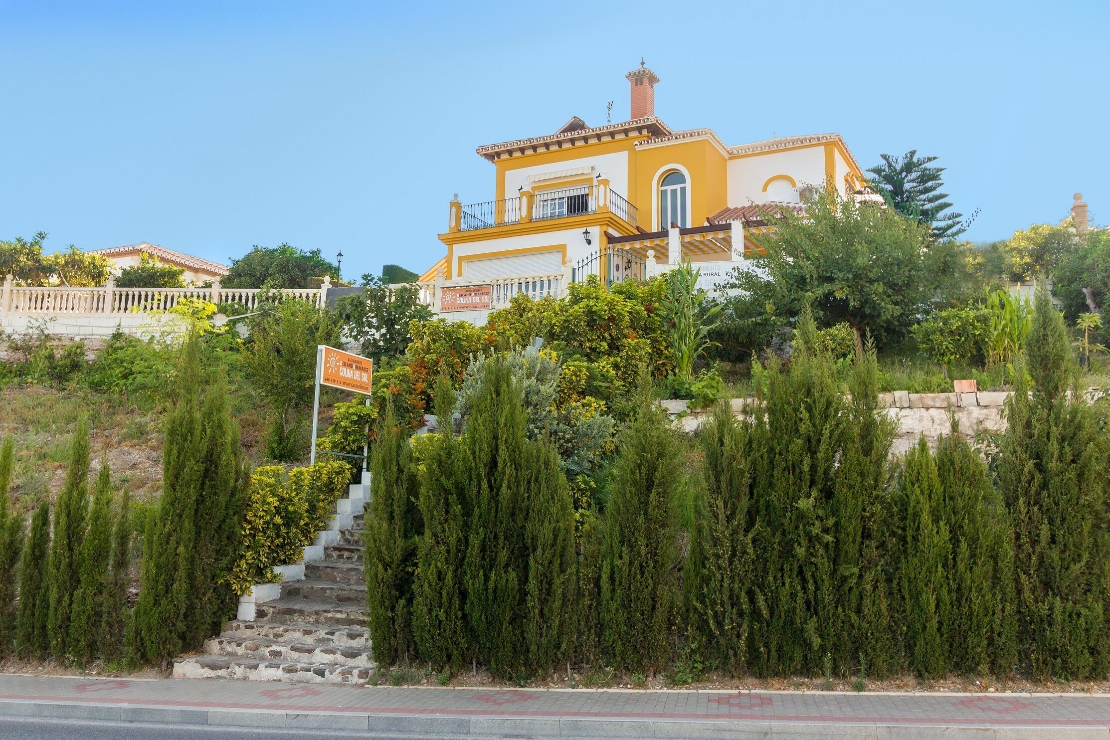 A yellow building with a balcony, surrounded by greenery and a stone wall.
