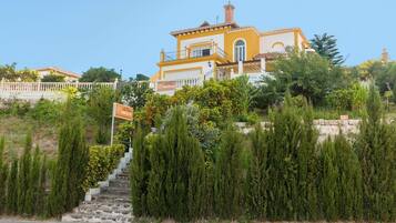 A yellow building with a balcony, surrounded by greenery and a stone wall.