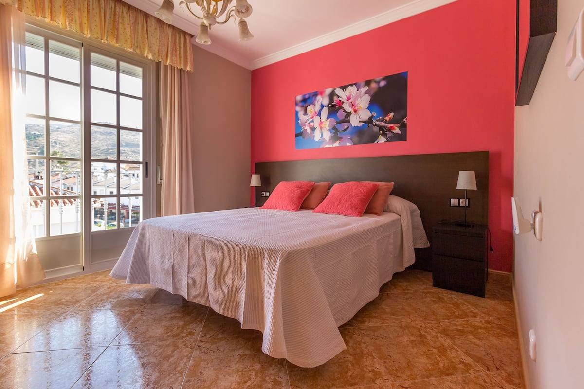 A bedroom with a large bed, a window with curtains, a painting on the wall, and a nightstand with a lamp.