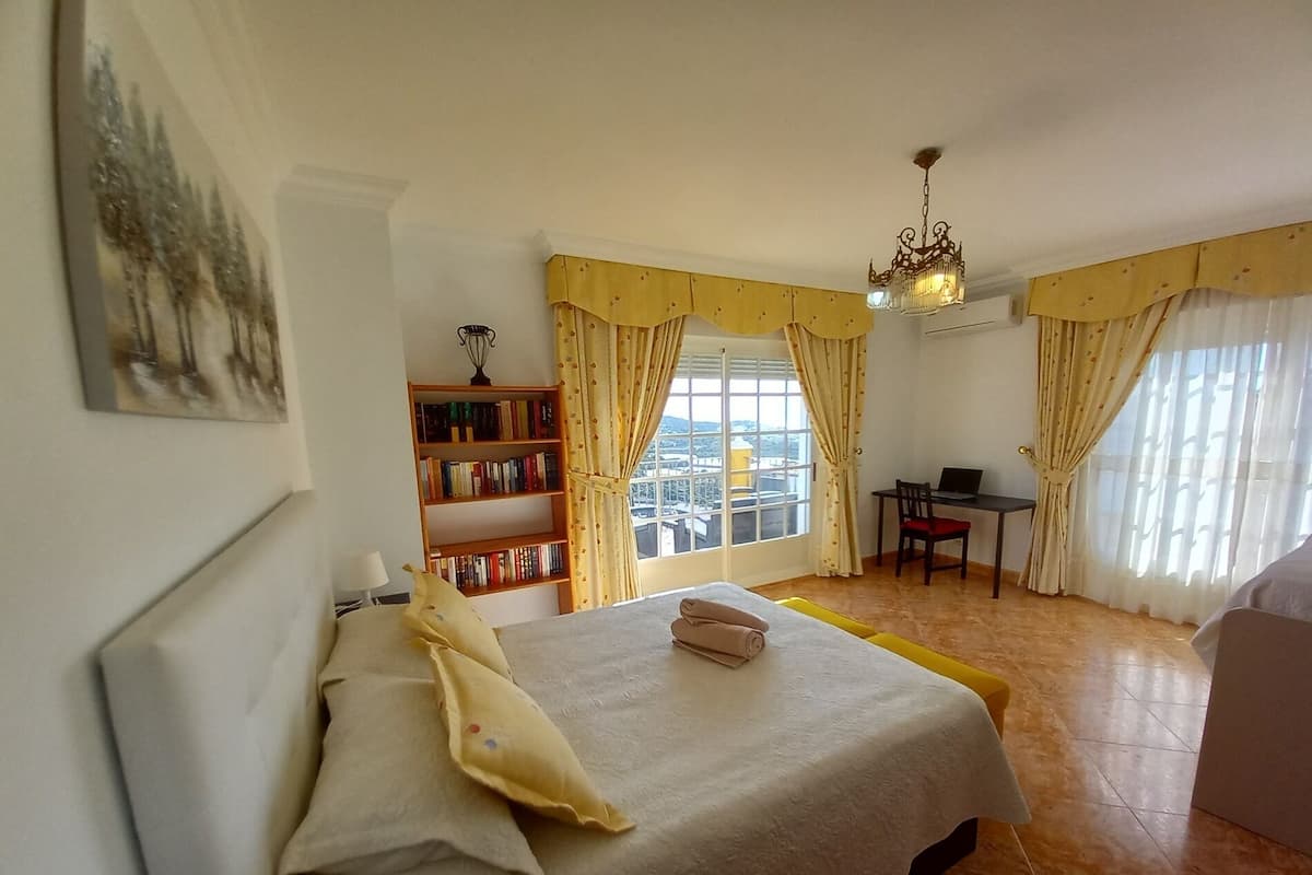 A hotel room with a bed, a desk with a laptop, a bookshelf, and a view of the city through large windows.