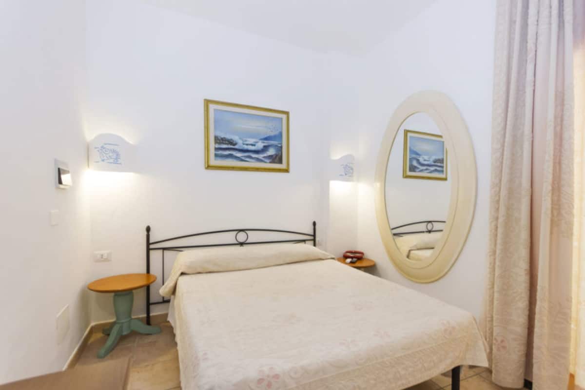 A bedroom with a bed, a round mirror, two wall-mounted lights, a small table, and two framed pictures.