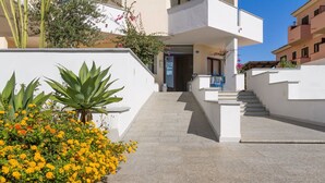 Ein modernes Gebäude mit weißer Fassade, einer Treppe zu einer Tür und einem Garten mit gelben Blumen und einer großen Pflanze.