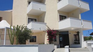 Ein mehrstöckiges Gebäude mit Balkonen, einem zentralen Eingang und einer kleinen Gartenanlage mit Kakteen und Bougainvillea.