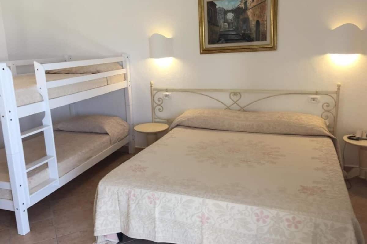 A bunk bed with a lower bunk and a single bed, both with floral bedspreads. There are two bedside tables with lamps, and a framed painting on the wall above the bed.