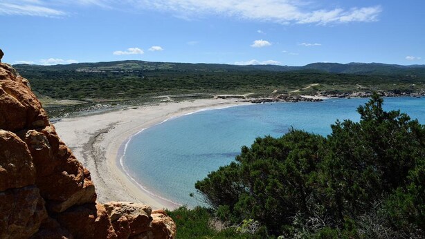Eine Küstenlandschaft mit Sandstrand, klarem, blauem Wasser und grünen Hügeln im Hintergrund.